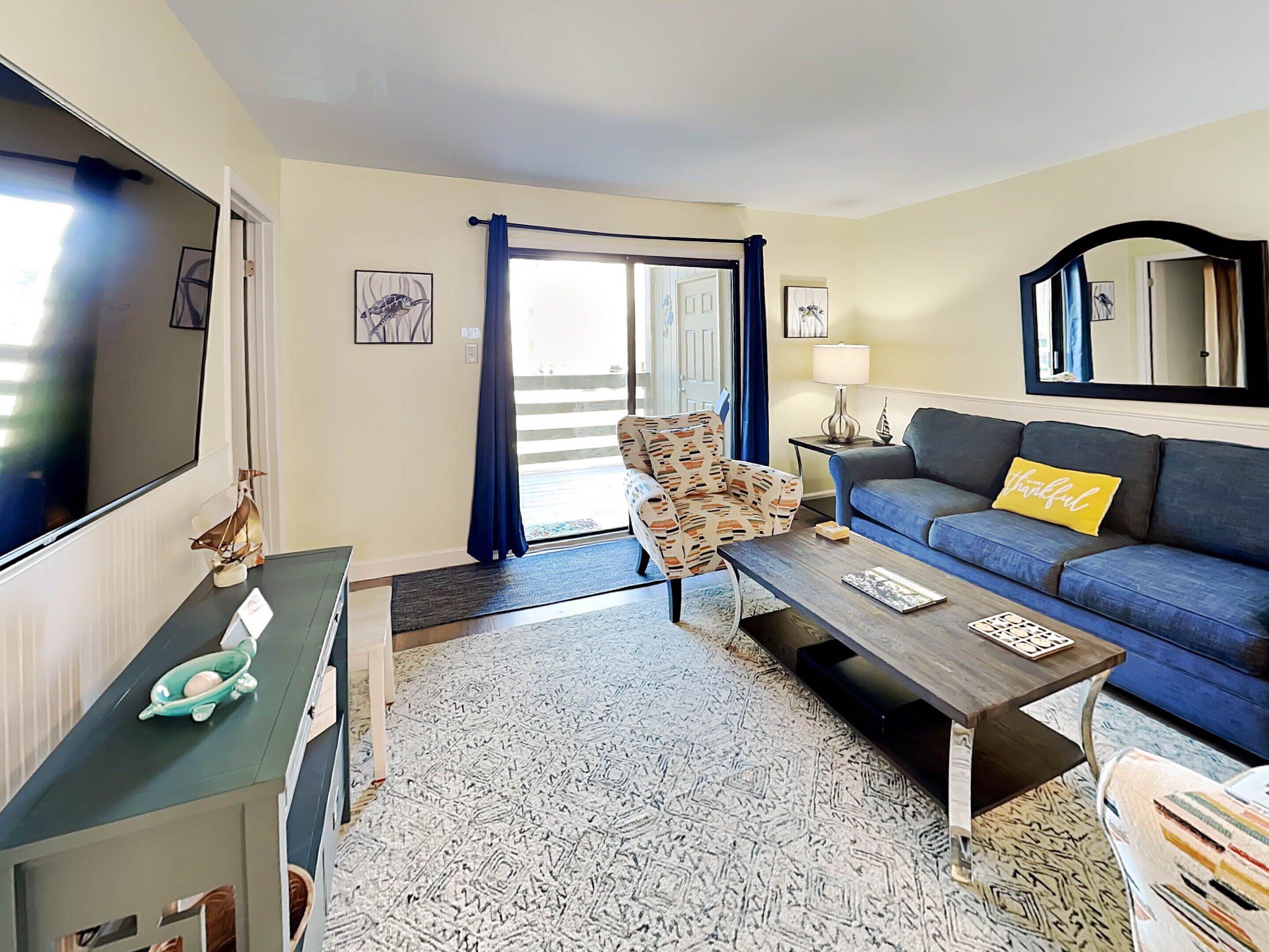 A living room with a sofa, a patterned armchair, a coffee table, a TV, and a door leading to a balcony.