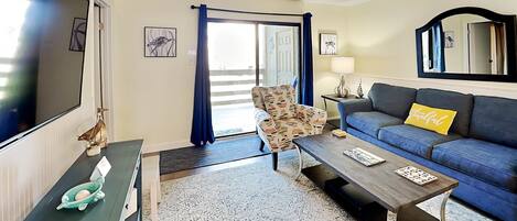 A living room with a sofa, a patterned armchair, a coffee table, a TV, and a door leading to a balcony.