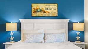 A bedroom with a blue wall, a white headboard, and a beach-themed welcome sign. - Lighthouse Point 33A by Tybee Vacation Rentals (Tybee Island)