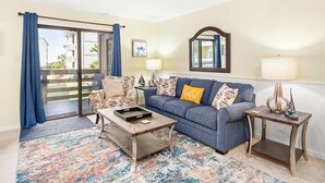 A living room with a blue sofa, a coffee table, a patterned rug, a side table with a lamp, and a view through a glass door. - Lighthouse Point 33A by Tybee Vacation Rentals (Tybee Island)