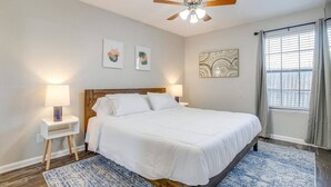 Une chambre à coucher comprenant un lit, un ventilateur de plafond, deux tables de chevet avec des lampes, une fenêtre avec des rideaux et des œuvres d’art accrochées aux murs.