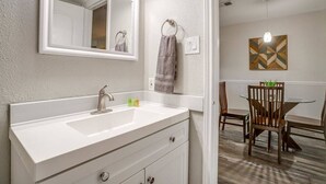Une salle de bain avec un lavabo blanc, un miroir et un porte-serviettes. On aperçoit, au fond, un coin repas avec une table et des chaises.
