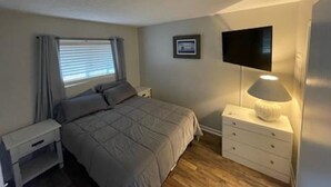 Une chambre à coucher comprenant un lit, une table de chevet, une commode, une lampe, un téléviseur et une fenêtre avec des rideaux.