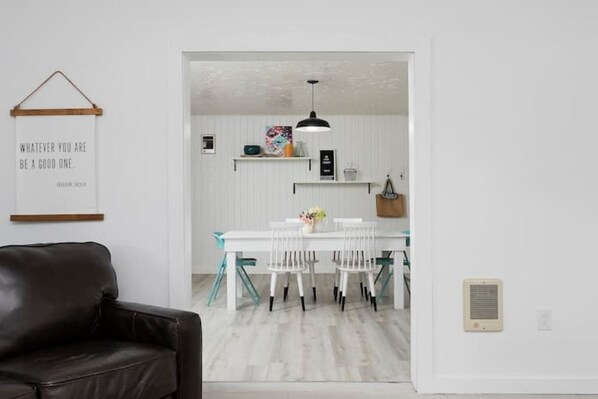 Una sala de estar moderna con un sofá de cuero marrón, una mesa de comedor blanca con cuatro sillas y una cocina con armarios blancos y una lámpara colgante.