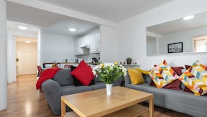 Una sala moderna con un sofá gris, una mesa de centro de madera y un jarrón de flores. Se aprecia una cocina al fondo, con armarios blancos y una manta roja sobre el sofá.