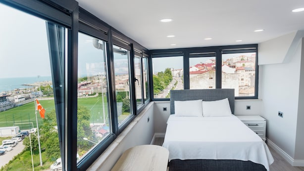 A modern bedroom with a large bed, a small table, and a view of the city through large windows. - Grand Family Home (Trabzon)