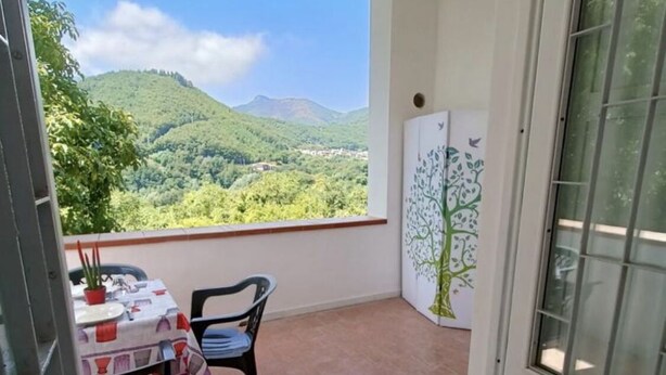 A balcony with a table and chairs, a view of green hills, and a door with a tree mural. - A cozy refuge in the sky (tramonti)