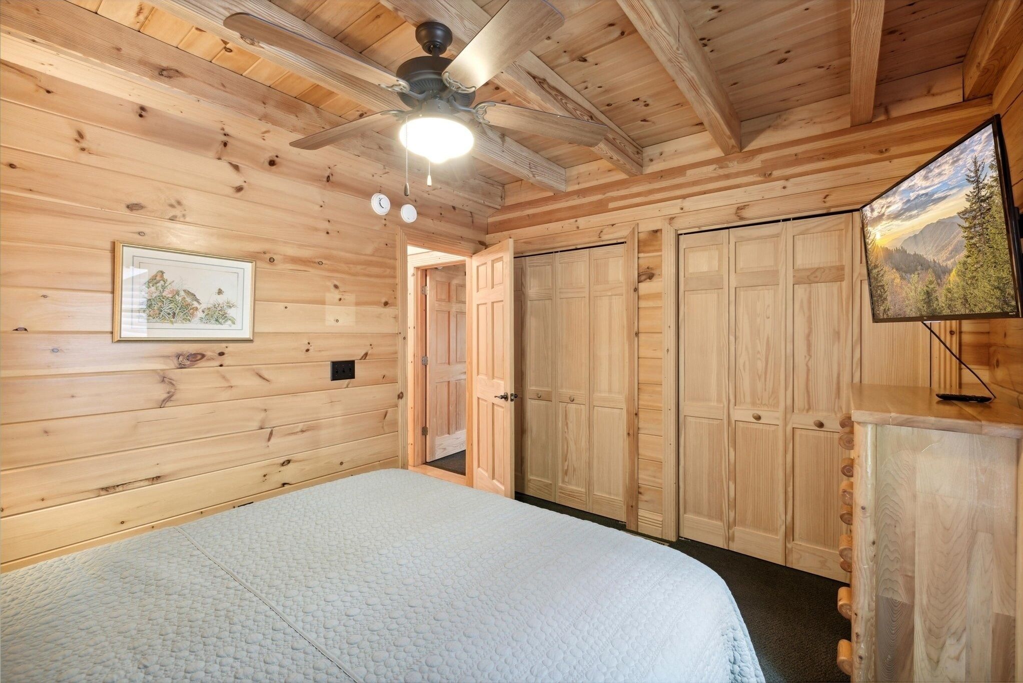 Une chambre dans une cabane en bois, avec un lit, un ventilateur de plafond, un téléviseur fixé au mur et des armoires en bois.