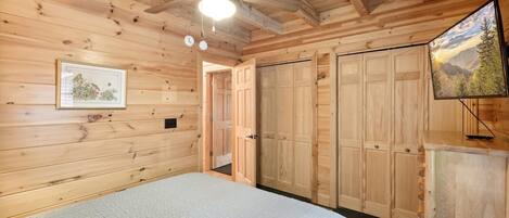 Une chambre dans une cabane en bois, avec un lit, un ventilateur de plafond, un téléviseur fixé au mur et des armoires en bois.