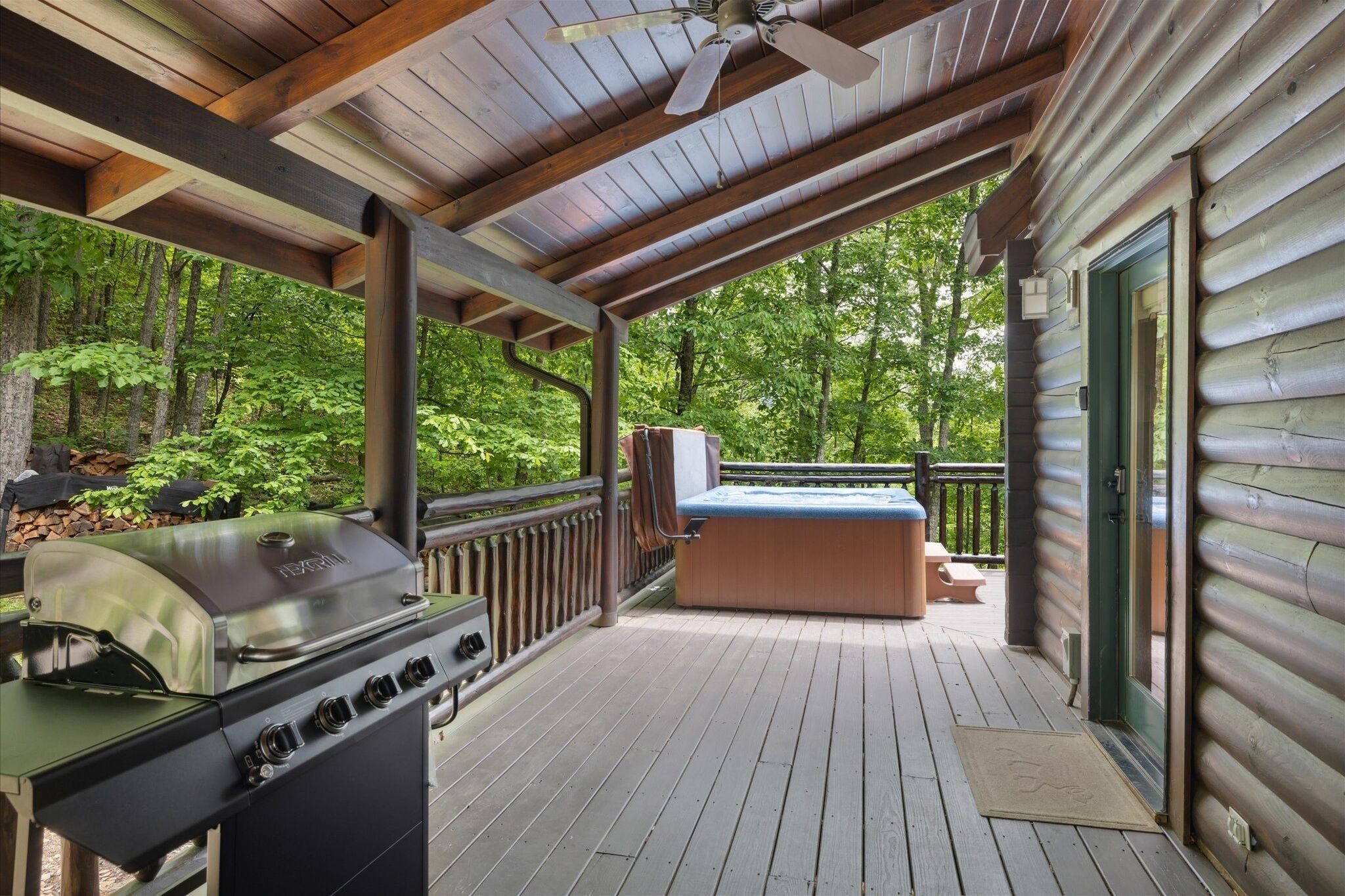 A covered deck with a grill, a hot tub, and a view of a forest.