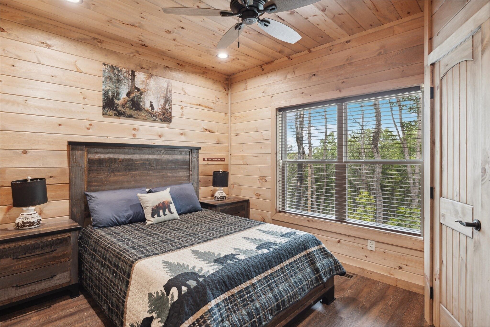 A wooden cabin bedroom with a bed, nightstands, a ceiling fan, and a large window with blinds.