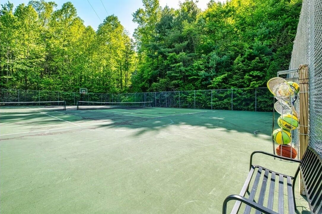 En udendørs tennisbane omgivet af træer, med en bænk og tennisraketter til siden.