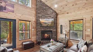 Interior acogedor de una cabaña con chimenea de piedra, paredes de madera y ventanas amplias con vistas al exterior.