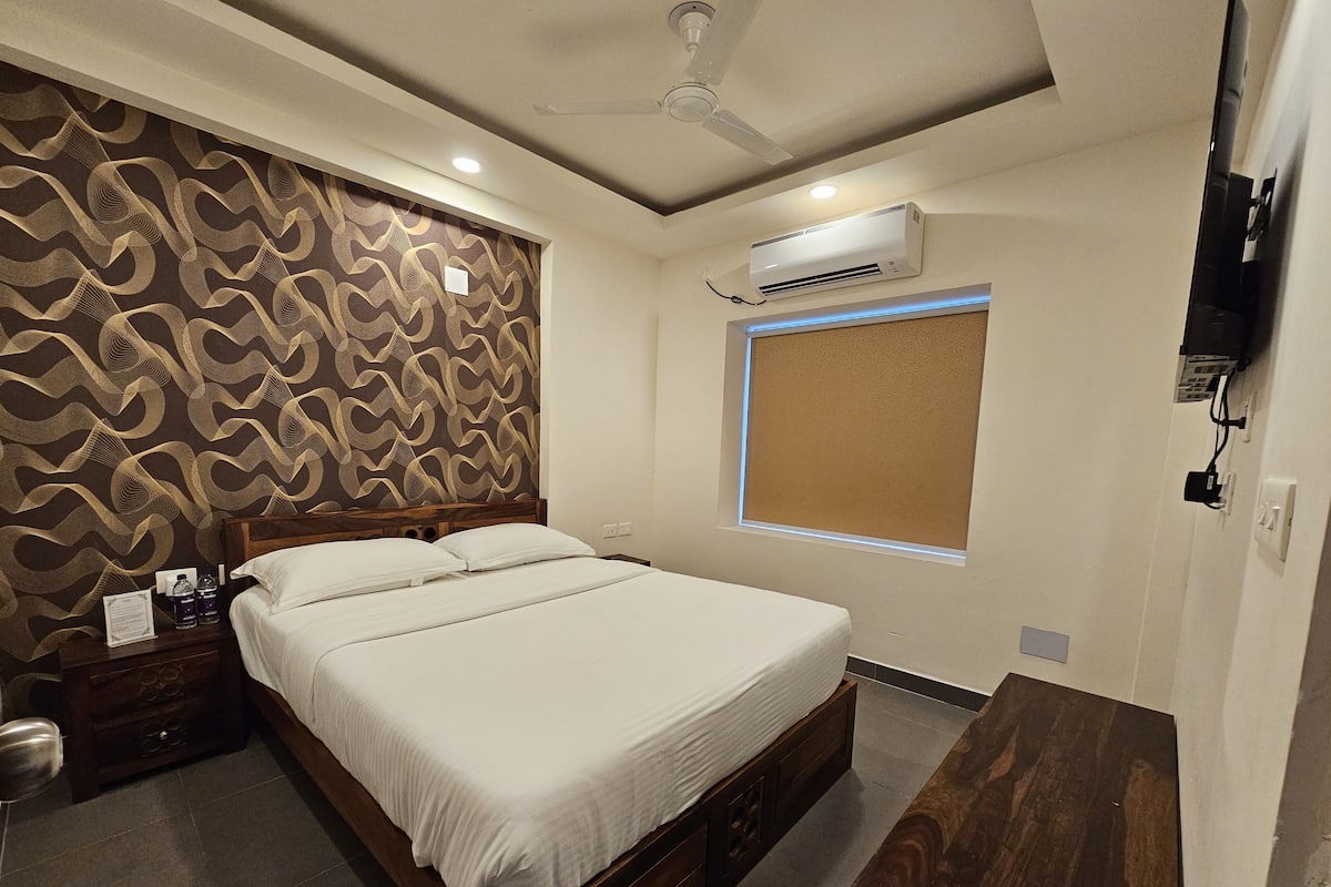 Habitación de hotel con cama, mesita de noche de madera, escritorio, ventilador de techo, aire acondicionado y ventana con persianas.