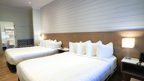 A hotel room with two beds, a wooden headboard, a bedside table with a lamp, and a bathroom visible in the background. - The Colonial Hotel - Premium Double Queen (Grand Bend)