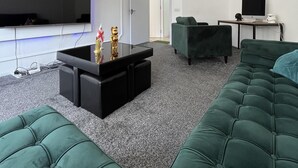 Una sala de estar moderna con un televisor de pantalla plana grande, una mesa baja oscura, sofás verdes y una mesita con una computadora y teclado.