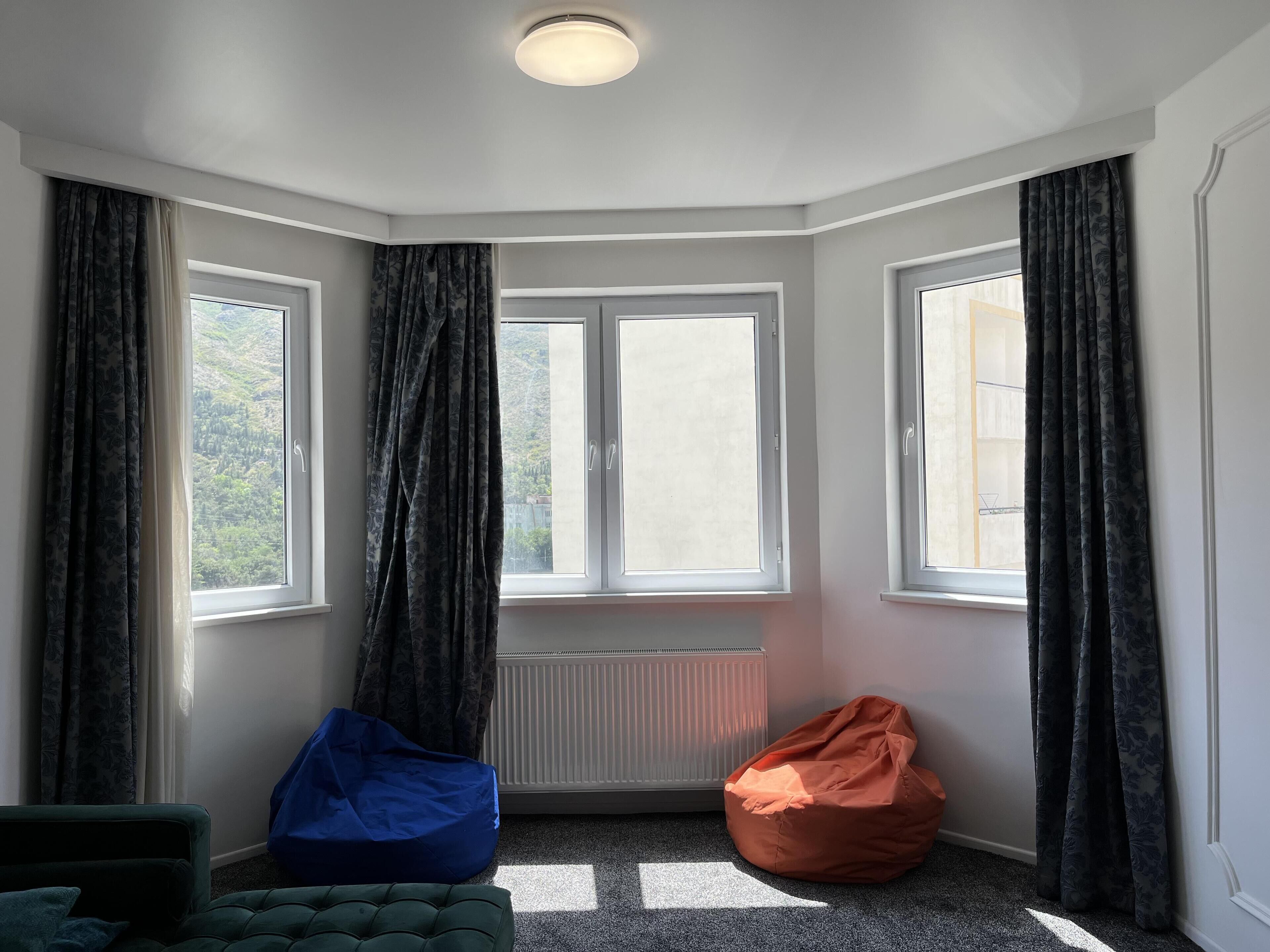Une pièce dotée de deux grandes fenêtres, d’un radiateur, d’un fauteuil poire bleu et d’un fauteuil poire orange.