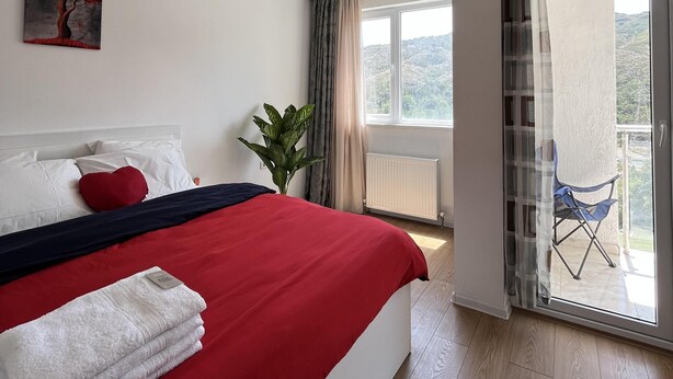 Un dormitorio con cama, una colcha roja y blanca, un cojín con forma de corazón rojo, una toalla blanca, una planta, un cuadro y vistas a un balcón con una silla.