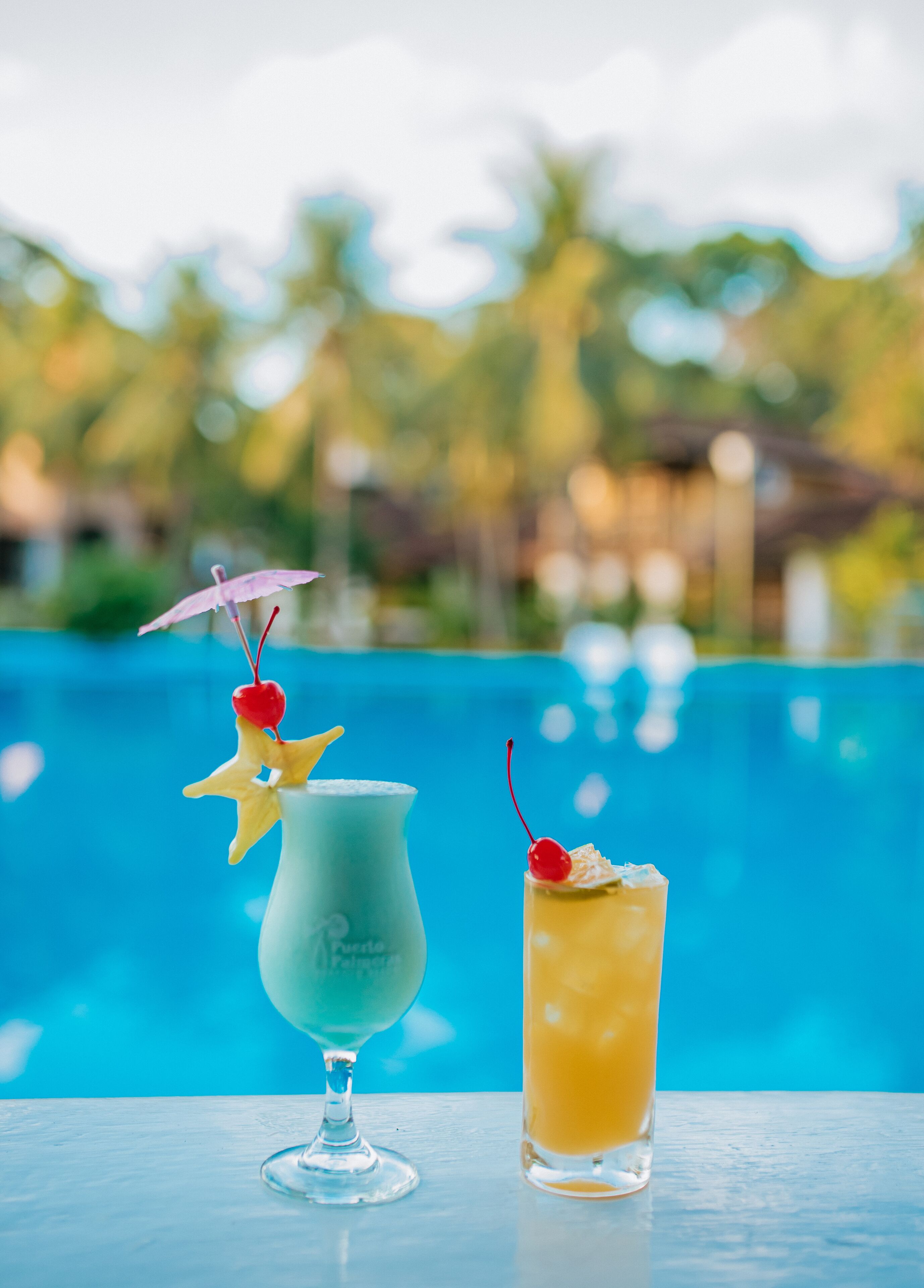 Dos còctels sobre una taula a la vora d'una piscina amb un paisatge tropical.