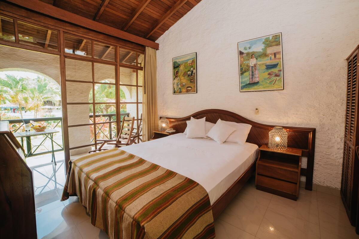 Une chambre à coucher avec un grand lit, une tête de lit en bois, deux tableaux accrochés au mur, une table de chevet avec une lampe, et une vue sur l’extérieur par une fenêtre.