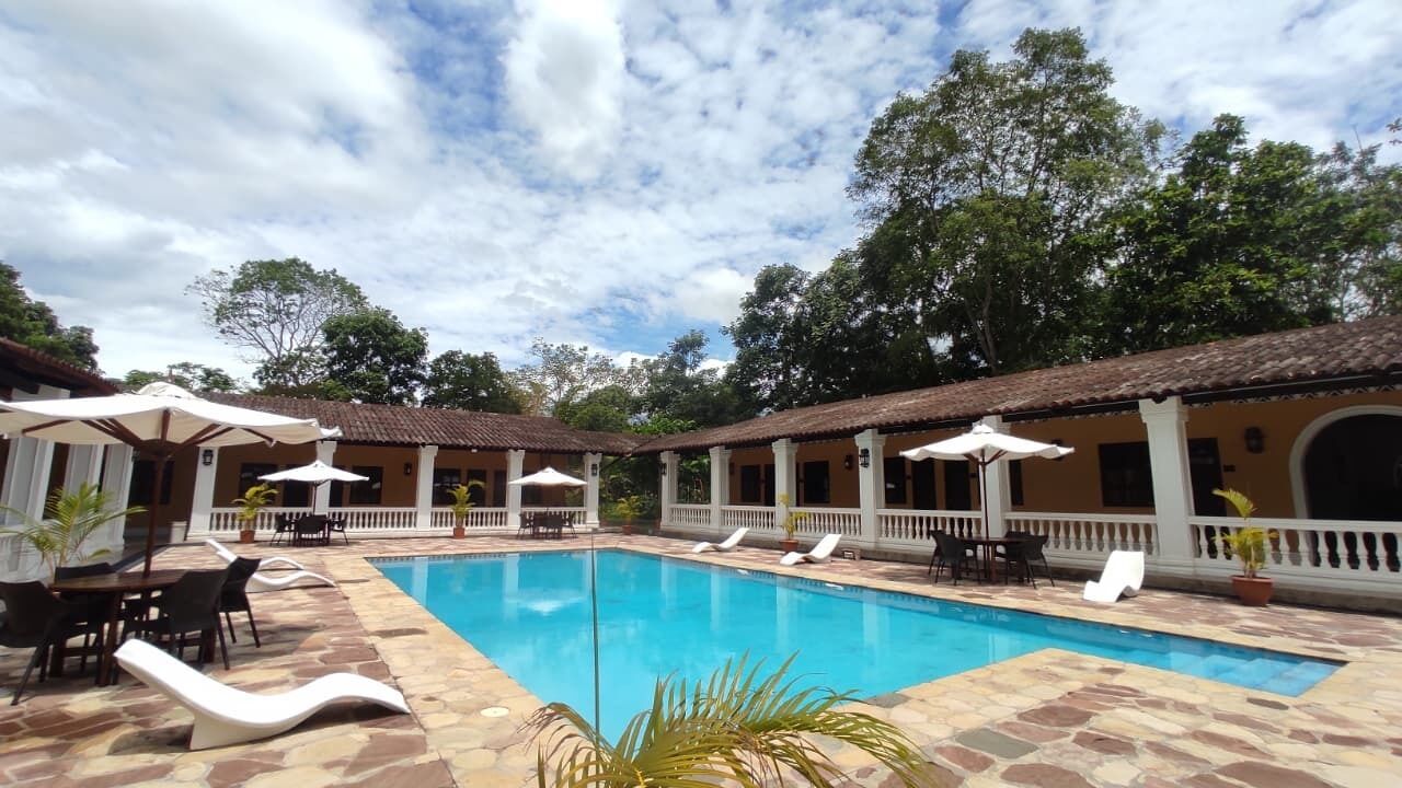 Una piscina con sillas de descanso y sombrillas, rodeada de zonas de estar cubiertas y plantas en macetas.