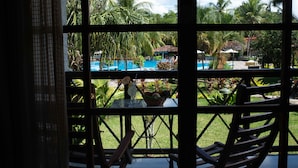 Une vue à travers une fenêtre sur un espace piscine, avec une table et des chaises, entouré de palmiers et sous un ciel dégagé.