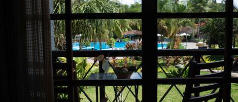 A view through a window of a pool area with a table and chairs, surrounded by palm trees and a clear sky.