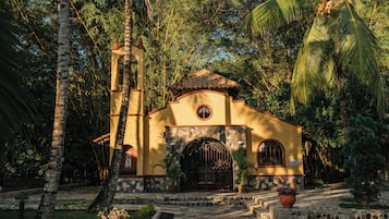 A small, yellow church with a stone facade and a bell tower, surrounded by lush greenery.