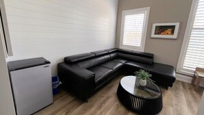 Een moderne woonkamer met een zwarte leren zithoek, een kleine koelkast, een ronde salontafel met een plant, en een lijst met een afbeelding aan de muur.