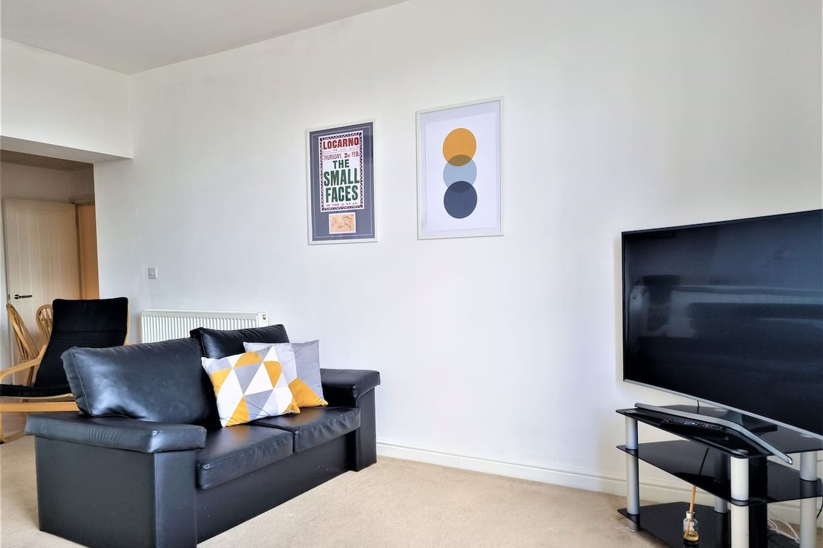 Een woonkamer met een zwarte bank, een flatscreen-tv en twee ingelijste posters aan de muur.