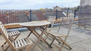 Une terrasse avec une table ronde et des chaises en bois, donnant sur un quartier résidentiel avec des maisons aux toits rouges et une église.