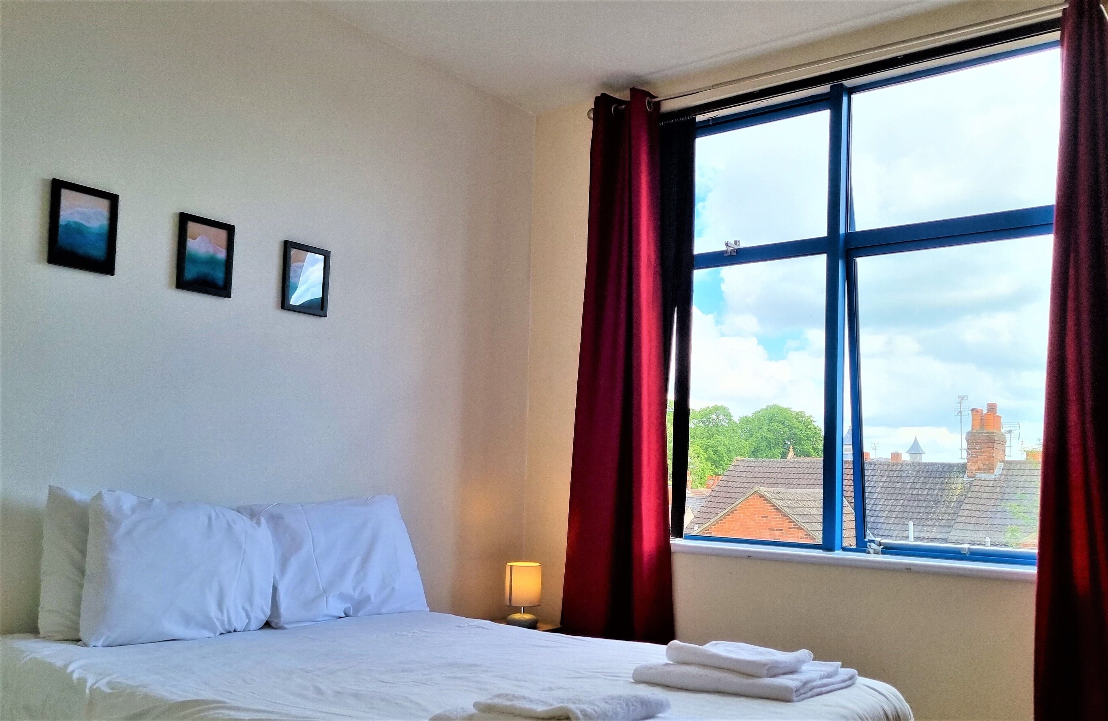 Une chambre d’hôtel avec un lit, une table de chevet, une fenêtre avec des rideaux et trois tableaux encadrés accrochés au mur.