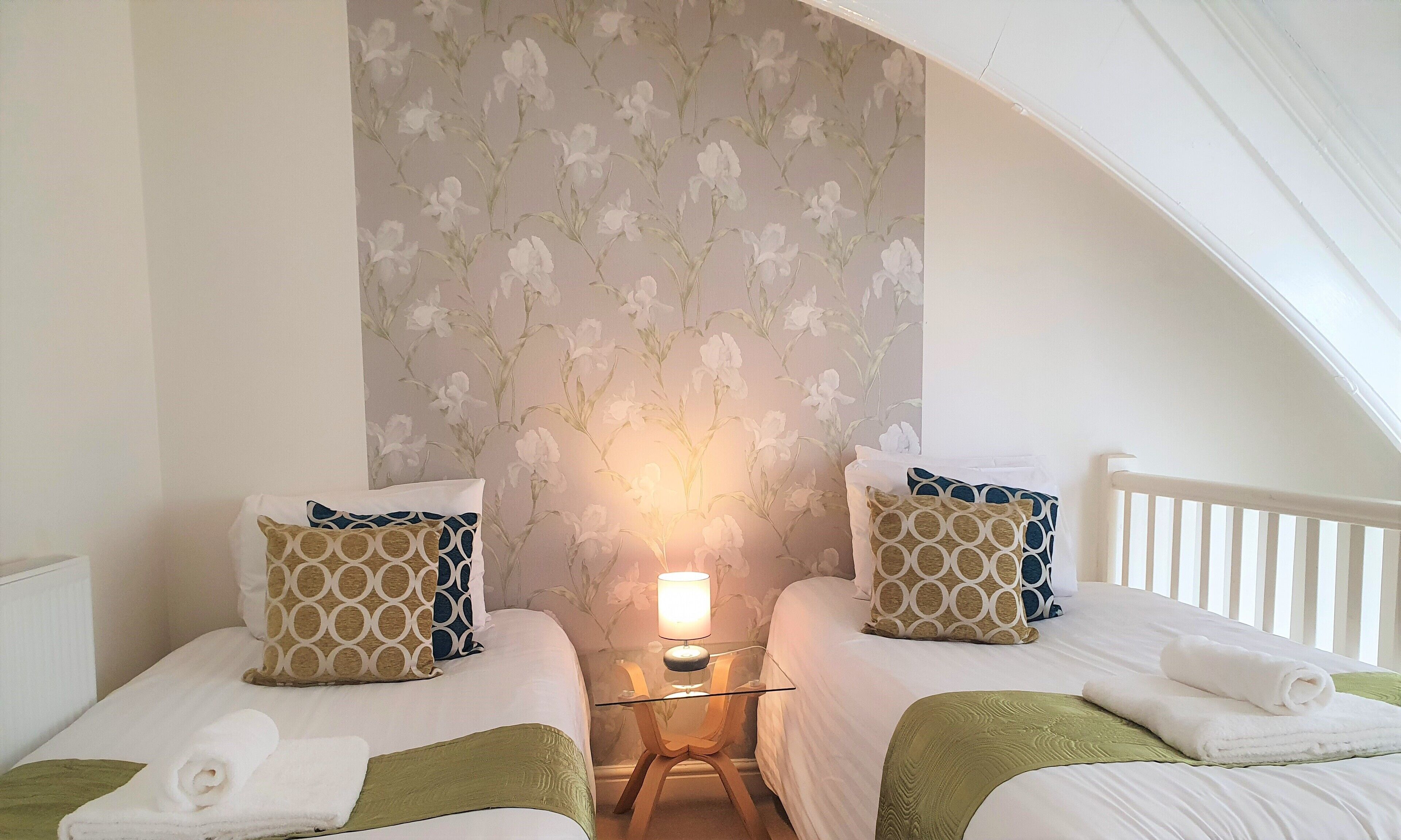 A bedroom with two beds, each with patterned pillows, a bedside table with a lamp, and a floral wallpaper backdrop.