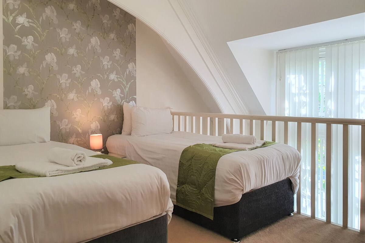 Een slaapkamer met twee bedden, een nachtkastje, een lamp en uitzicht op de omgeving door een raam met jaloezieën.