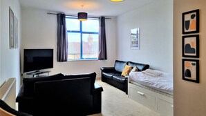 Une pièce moderne et bien éclairée, dotée d’une grande fenêtre, d’un canapé en cuir noir, d’un lit avec un matelas blanc, d’une télévision et d’un luminaire suspendu.