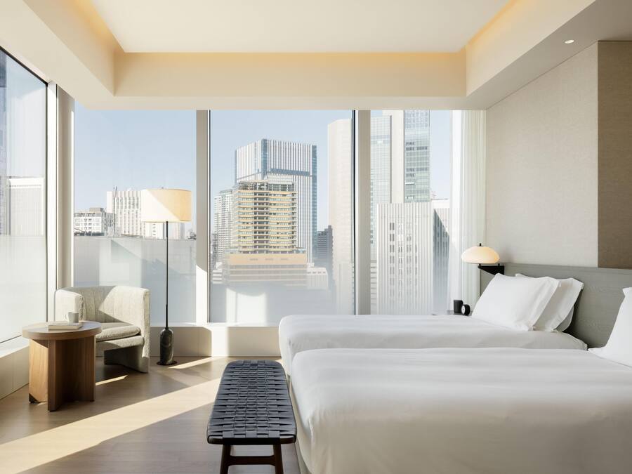 A modern hotel room with a large window, a bed, a chair, a small table, and a lamp.