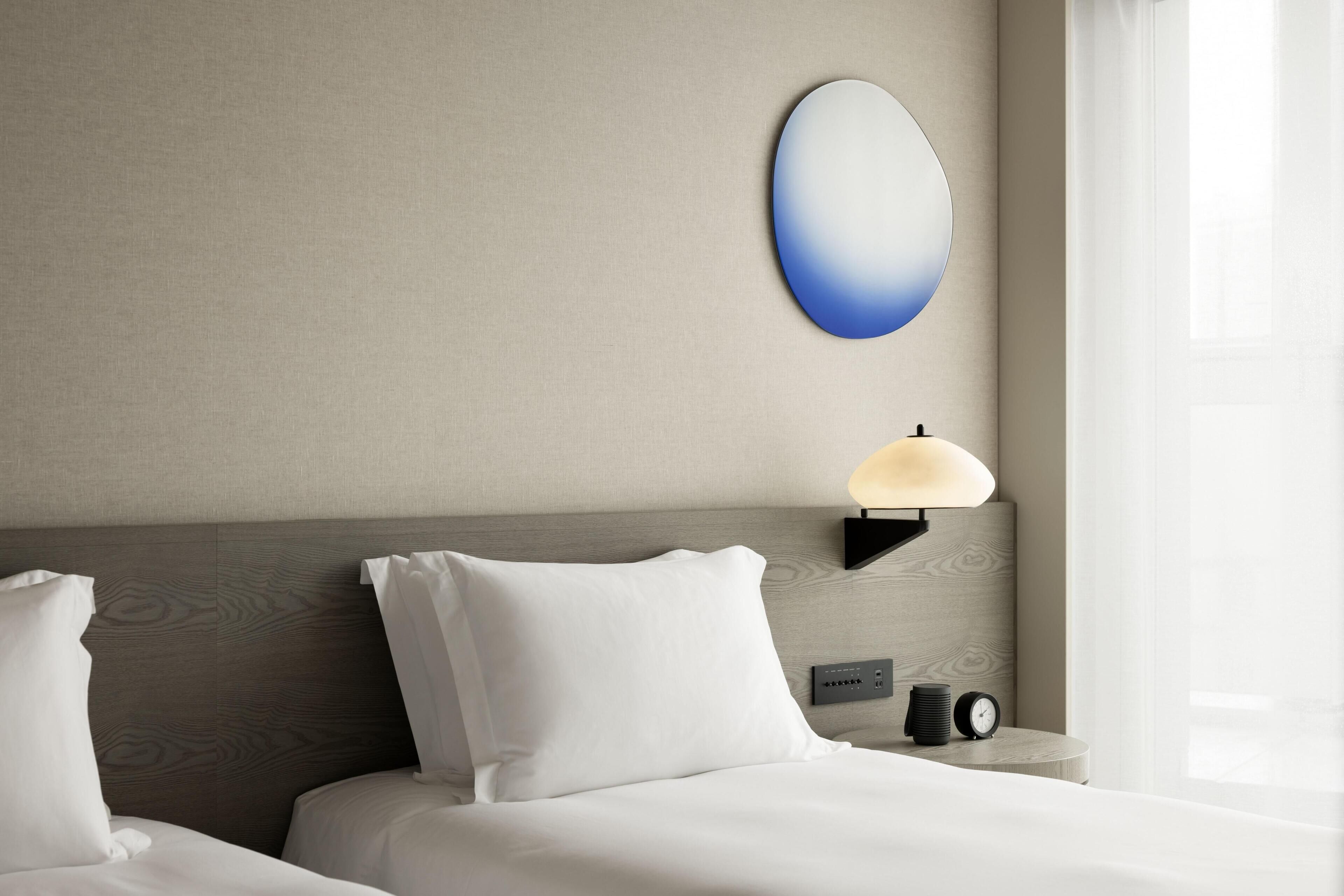 A hotel room with a bed, pillows, a wall-mounted lamp, a clock, and a window with curtains.