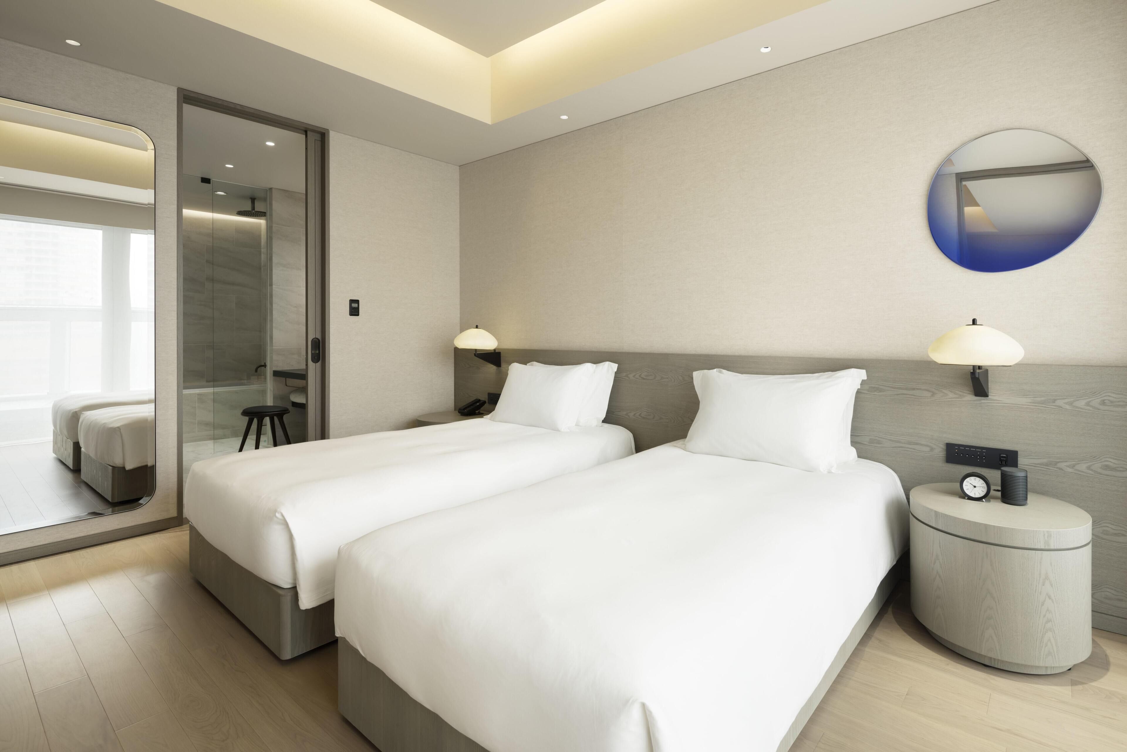 A hotel room with two beds, a round mirror, a nightstand with a clock, and a bathroom with a glass door.