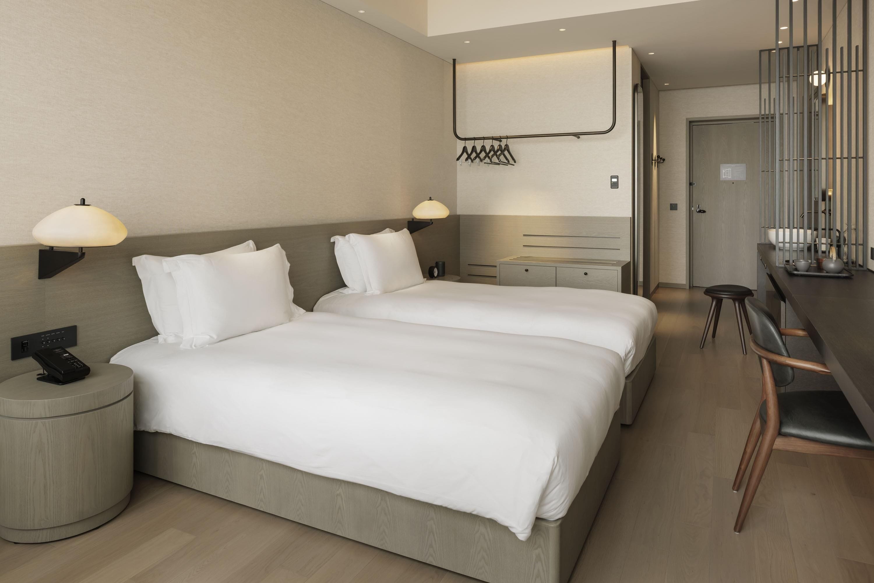 A hotel room with two beds, a desk with chairs, a small round table, a wall-mounted rack, and a door.
