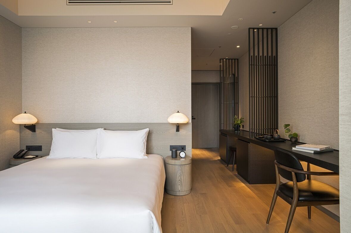 A modern hotel room with a large bed, bedside lamps, a desk with a chair, and a small side table.