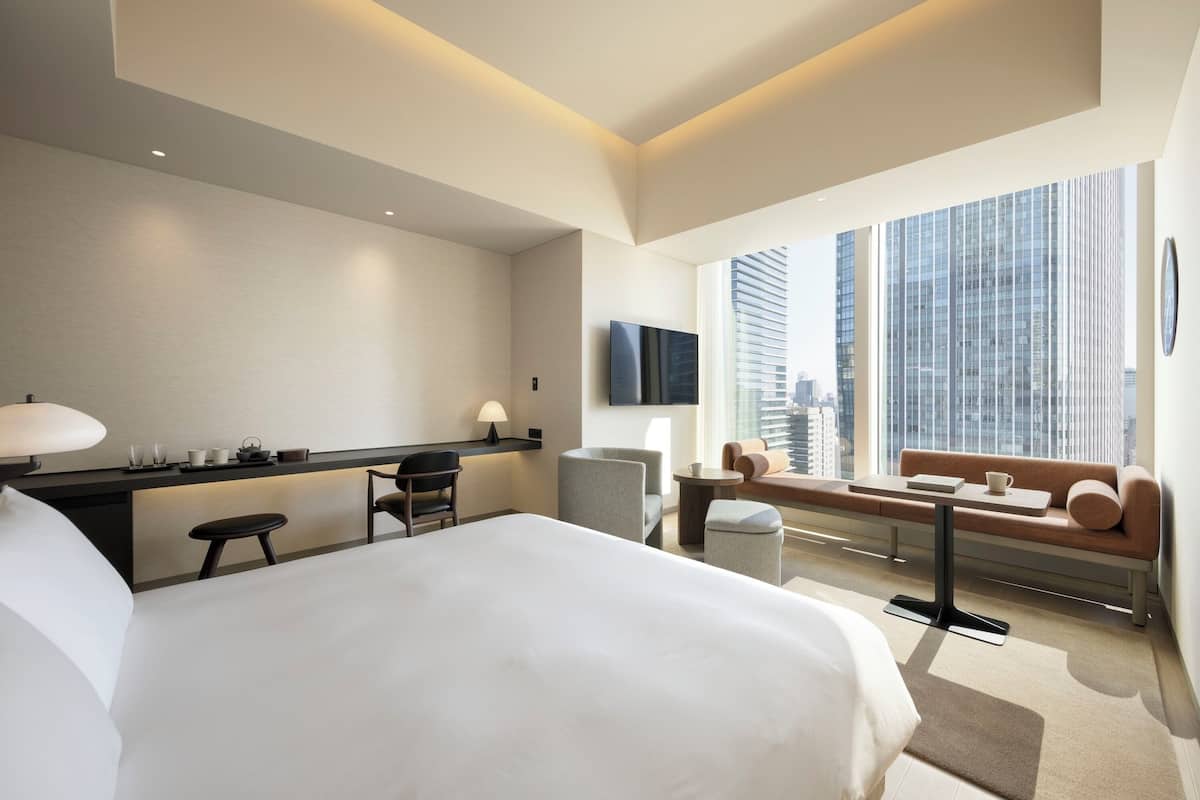 A modern hotel room with a large bed, a desk with a flat-screen TV, a sofa, and a window with a city view.
