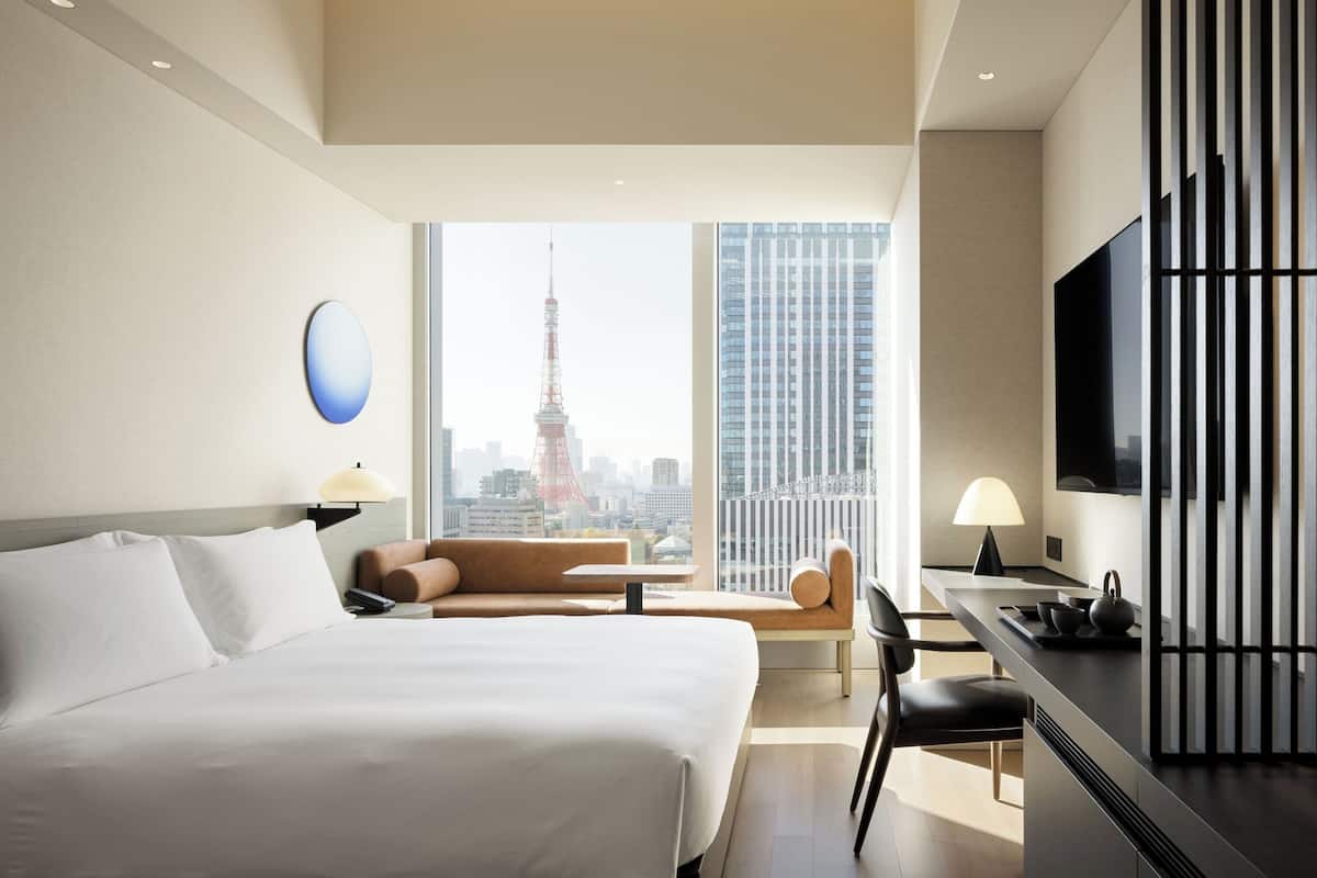A hotel room with a bed, a sofa, a desk, a TV, and a view of a cityscape including a tower.
