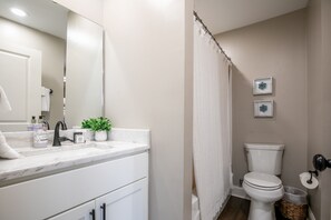 Ein Badezimmer mit Waschbecken, Spiegel und Toilette. An der Wand hängen zwei Bilder und auf dem Waschtisch steht ein Topf mit einer Pflanze.