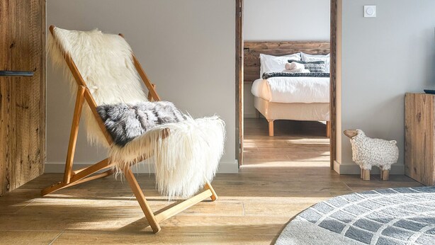 Een kamer met een houten vloer, een zachte witte fauteuil, een bed met een houten hoofdbord en een klein wit schaapsbeeldje.