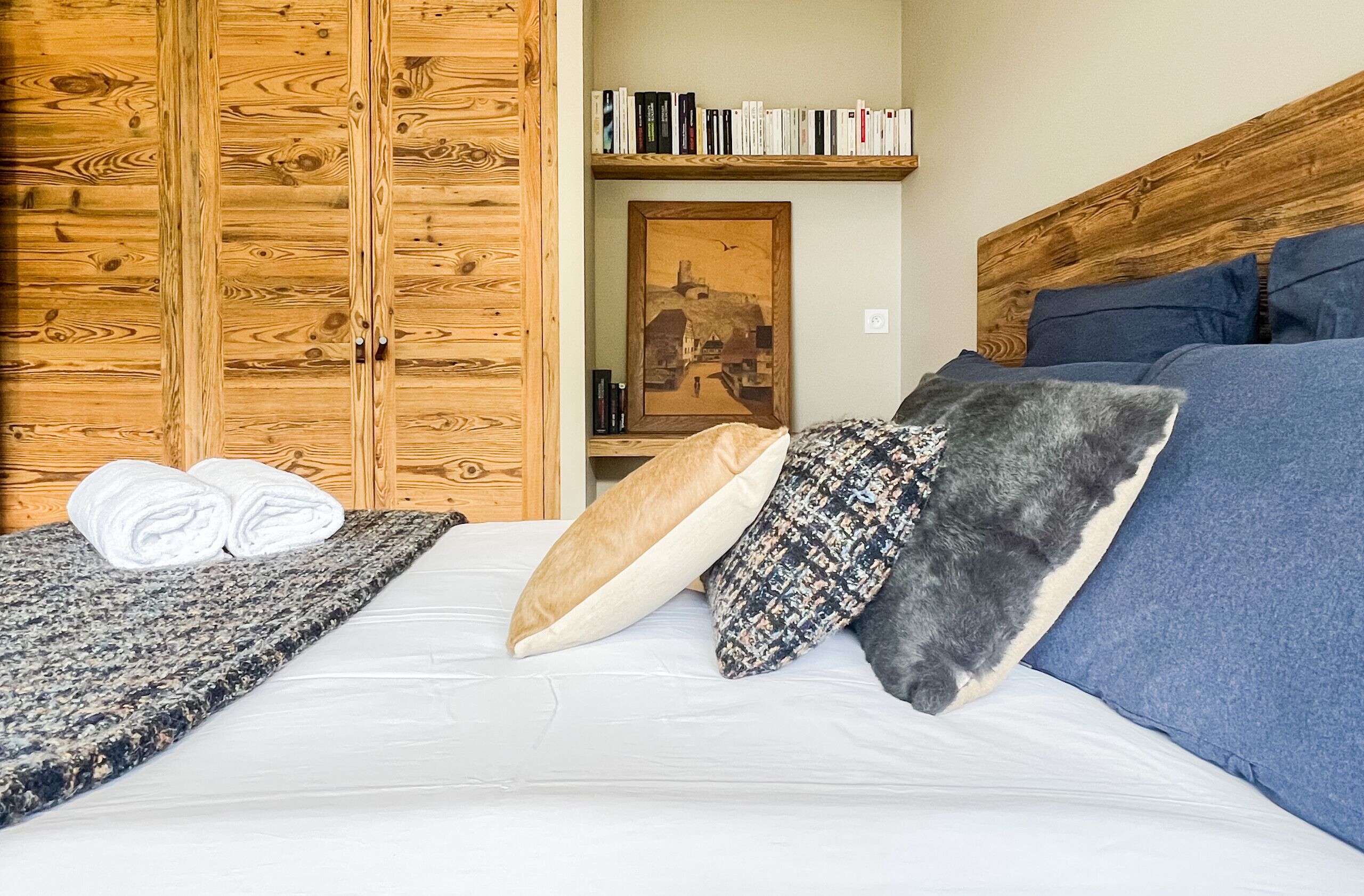 Un lit bien fait, recouvert d’une couette blanche, avec deux serviettes enroulées et trois oreillers. Une armoire en bois se trouve à gauche, et une bibliothèque remplie de livres est placée au-dessus du lit.