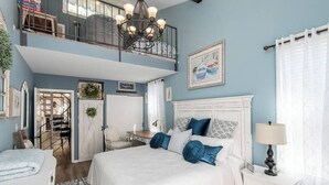 A bedroom with a large bed, a chandelier, a white nightstand, and a view of the outside. - Bayside Hideaway, Williams Bay, WI (Williams Bay)
