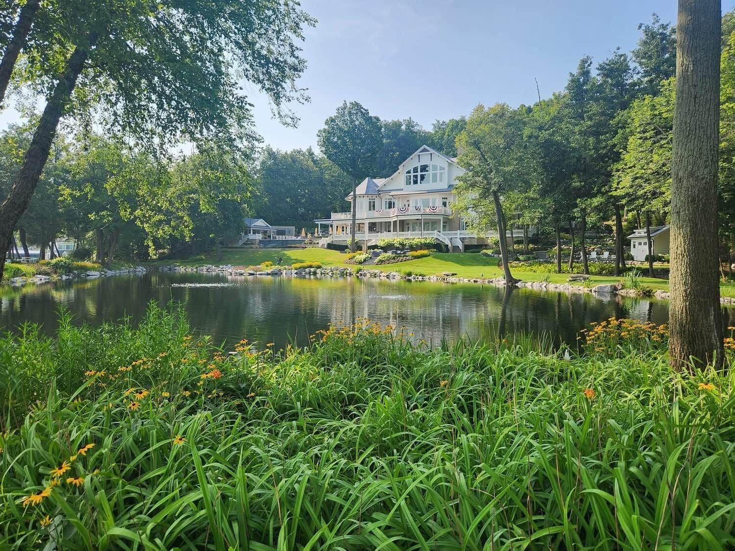 Une grande maison avec un étang devant, entourée de verdure et d’arbres.