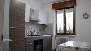 Una cocina compacta con estufa, horno y gabinetes blancos. Hay una ventana con cortinas y un reloj en la pared.