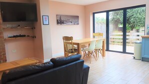 A living room with a black leather sofa, a wooden dining table with chairs, a TV mounted on the wall, and a large window with a view of greenery. - Large 5 Bedroom House Terrace Parking (Swindon)