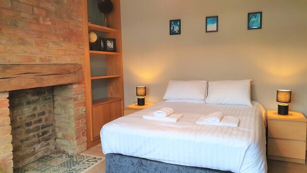 A neatly made bed with white linens and two pillows, a wooden nightstand with a lamp, a brick fireplace, and a shelf with decorative items. - Large 5 Bedroom House Terrace Parking (Swindon)